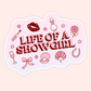 Pink and red retro-inspired vinyl sticker featuring the phrase “Life of a Showgirl” surrounded by glamorous icons like pearls, bows, lipstick, and champagne.