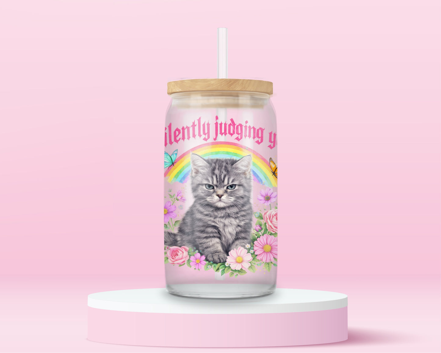 Glass can tumbler with bamboo lid and glass straw featuring a grumpy grey kitten, rainbow, flowers, butterflies, and pink text that says Silently Judging You on a pink background.
