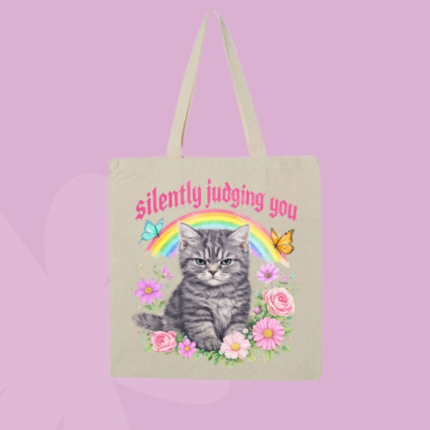 Beige canvas tote bag featuring a grumpy grey kitten, rainbow, flowers, butterflies, and pink text that says Silently Judging You.