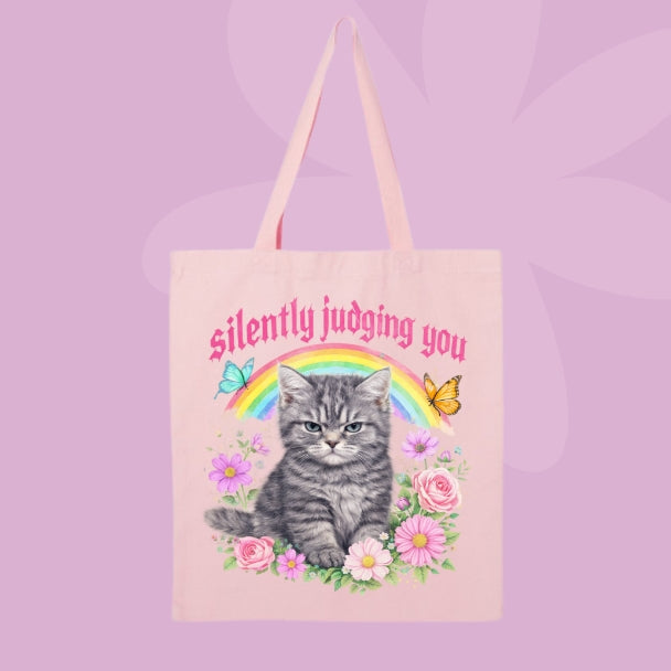 Pink canvas tote bag featuring a grumpy grey kitten, rainbow, flowers, butterflies, and pink text that says Silently Judging You.