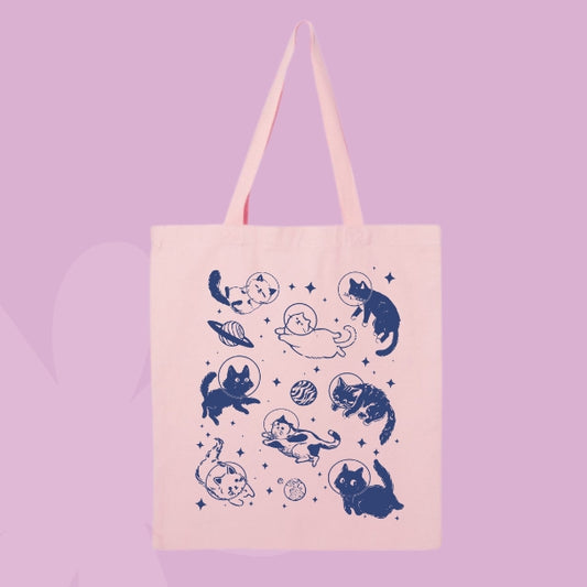 Pink tote bag with a navy design of cats floating in space with planets, stars, and astronaut helmets.