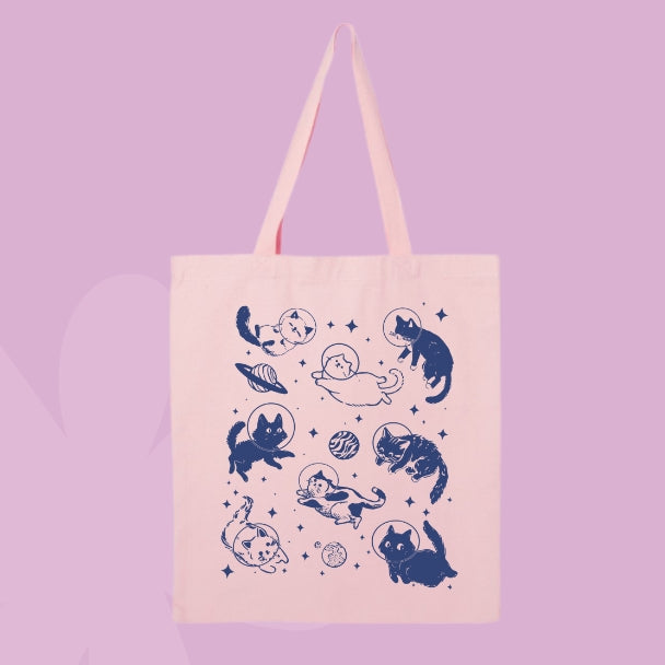 Pink tote bag with a navy design of cats floating in space with planets, stars, and astronaut helmets.