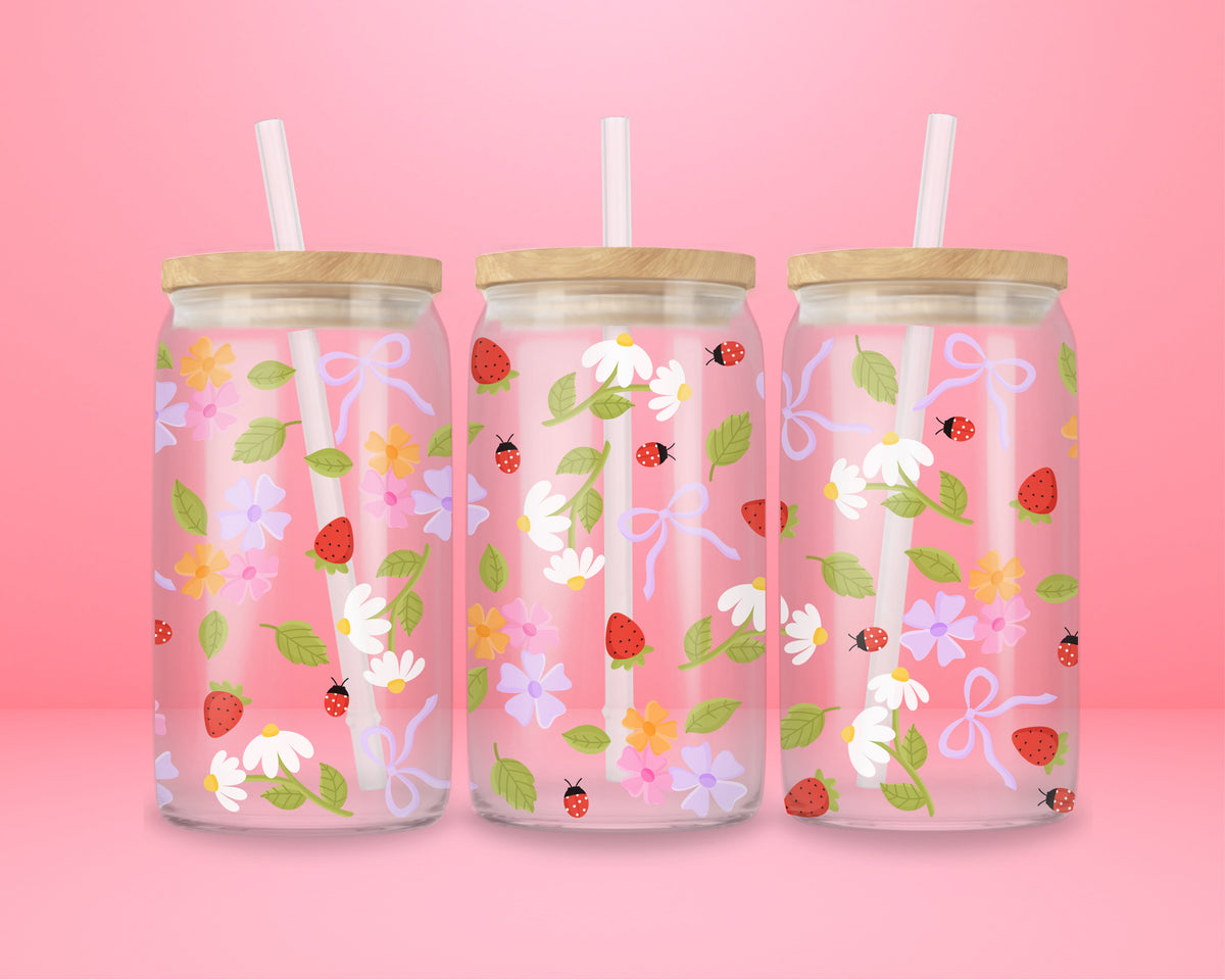 Glass can tumbler with bamboo lid and glass straw featuring strawberries, daisies, pastel flowers, green leaves, ladybugs, and lavender bow details on a pink background.