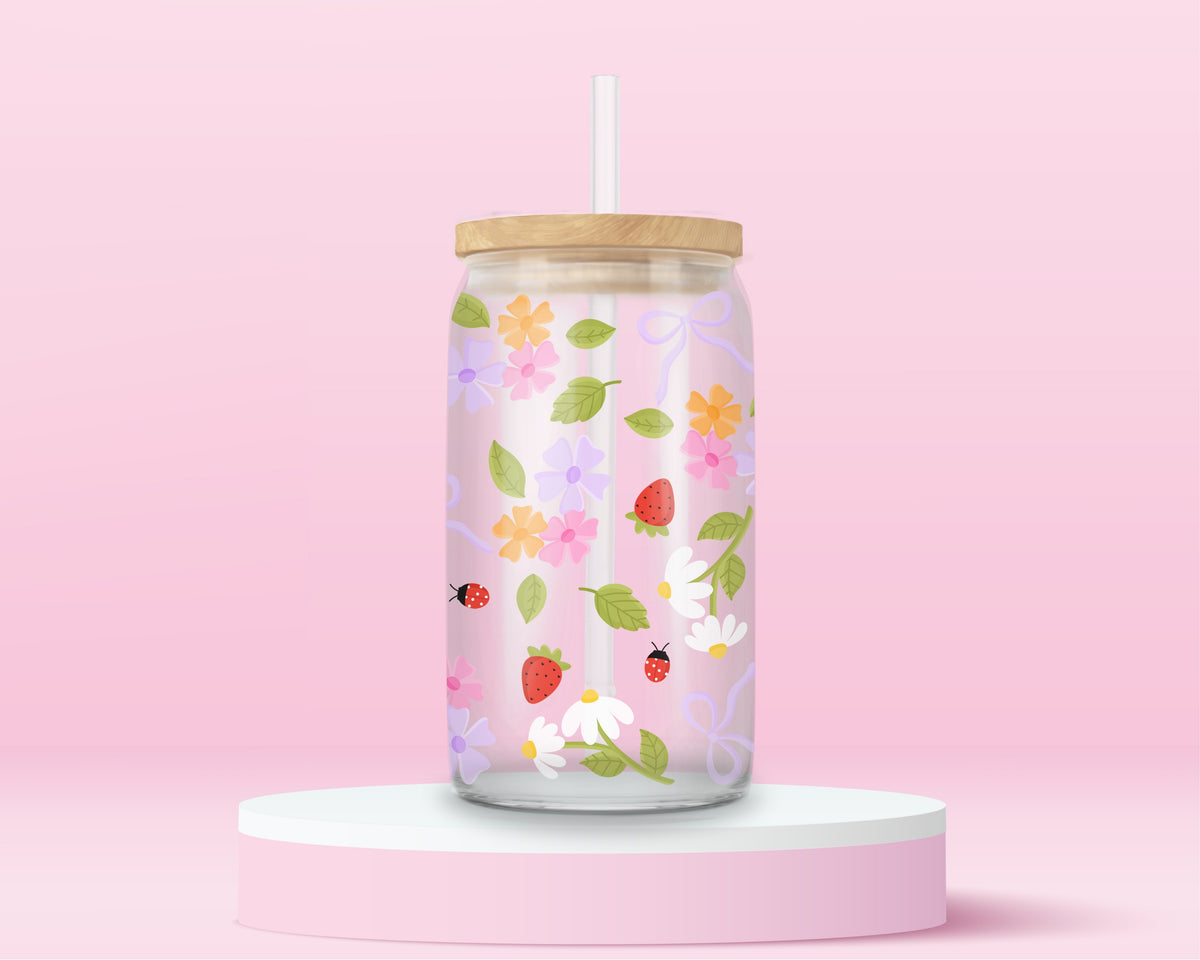 Glass can tumbler with bamboo lid and glass straw featuring strawberries, daisies, pastel flowers, green leaves, ladybugs, and lavender bow details on a pink background.