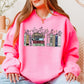 A woman wearing a bright pink Taylor Swift-themed crewneck sweater featuring illustrated book spines labeled with each Taylor Swift album from Debut to The Life of a Showgirl, with a cat resting on top and flowers blooming between the books.
