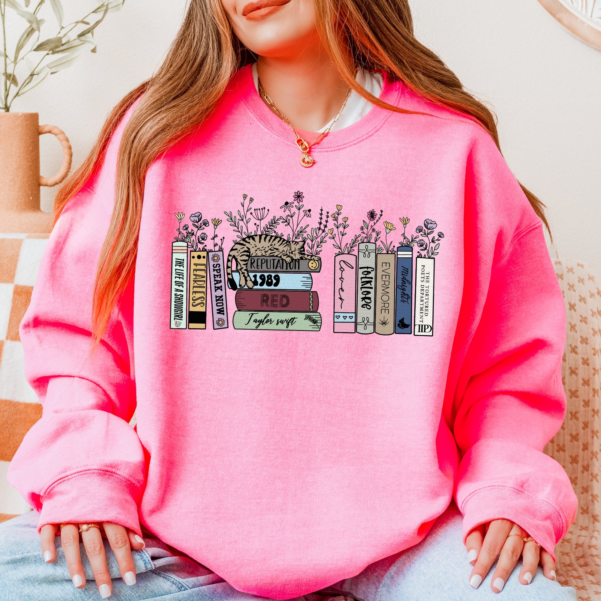 A woman wearing a bright pink Taylor Swift-themed crewneck sweater featuring illustrated book spines labeled with each Taylor Swift album from Debut to The Life of a Showgirl, with a cat resting on top and flowers blooming between the books.