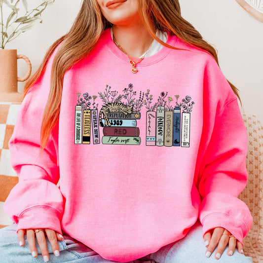 A woman wearing a bright pink Taylor Swift-themed crewneck sweater featuring illustrated book spines labeled with each Taylor Swift album from Debut to The Life of a Showgirl, with a cat resting on top and flowers blooming between the books.