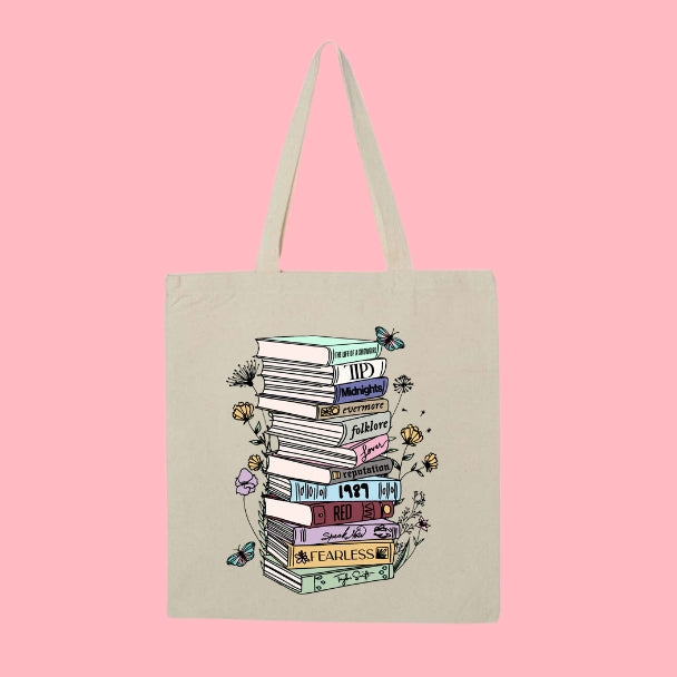 Beige tote bag with a tall stack of books labeled with Taylor Swift album names, surrounded by flowers and butterflies.