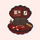 A deep red Polly Pocket–style seashell case opened to reveal a dramatic crime scene setting, featuring a dark sofa, crime files, caution tape, a candle, and thriller books labeled “Thriller.”