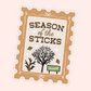 A die-cut sticker with a vintage postage stamp border and the text 'SEASON of the STICKS' in brown lettering. The design includes simple black line drawings of leafless trees, small brown leaves, white sparkles, and a green sign reading 'Vermont' on a pale pink background.