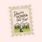 A die-cut postage stamp style sticker with the phrase “You’re GONNA GO Far” in brown script and serif lettering, above a simple landscape illustration of mountains, trees, grass, and a winding road. The stamp has a beige border with a white inner border and is shown on a pale pink background.