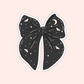 A die-cut sticker of a large black bow decorated with a celestial pattern of stars, moons, and small bats, shown on a solid pale pink background.