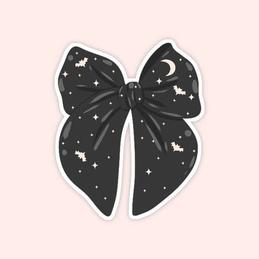 A die-cut sticker of a large black bow decorated with a celestial pattern of stars, moons, and small bats, shown on a solid pale pink background.