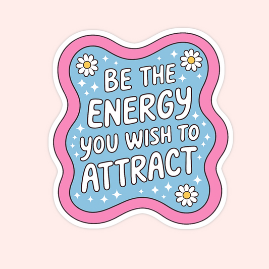 A die-cut sticker with a wavy scalloped border, featuring the motivational quote 'BE THE ENERGY YOU WISH TO ATTRACT' in white bubble letters on a light blue background with white daisies and sparkles. The sticker is presented on a solid pale pink background.
