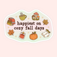 A die-cut sticker with a wavy border featuring autumn illustrations and the text 'happiest on cozy fall days' in dark brown lettering, shown on a pale pink background.