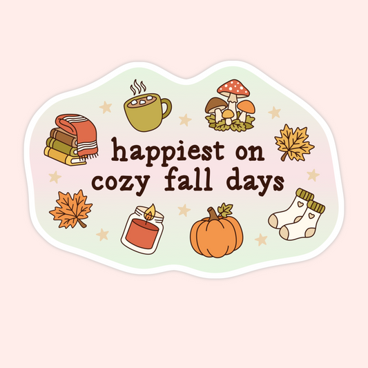 A die-cut sticker with a wavy border featuring autumn illustrations and the text 'happiest on cozy fall days' in dark brown lettering, shown on a pale pink background.