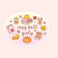 An oval sticker or decal with a white border, featuring a cozy fall illustration with the words 'cozy fall girly' in brown script. The design includes a candle, stack of books, coffee cup, book, slice of pie, pumpkins, leaves, flowers, and striped socks on a pale yellow-to-pink gradient background, set against a light peach background.