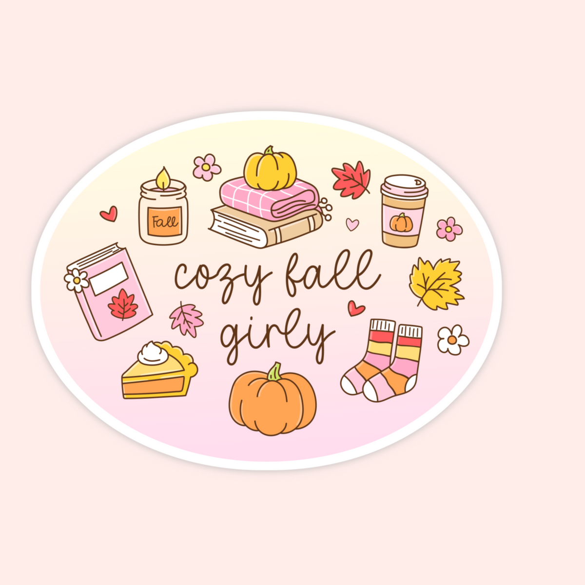 An oval sticker or decal with a white border, featuring a cozy fall illustration with the words 'cozy fall girly' in brown script. The design includes a candle, stack of books, coffee cup, book, slice of pie, pumpkins, leaves, flowers, and striped socks on a pale yellow-to-pink gradient background, set against a light peach background.