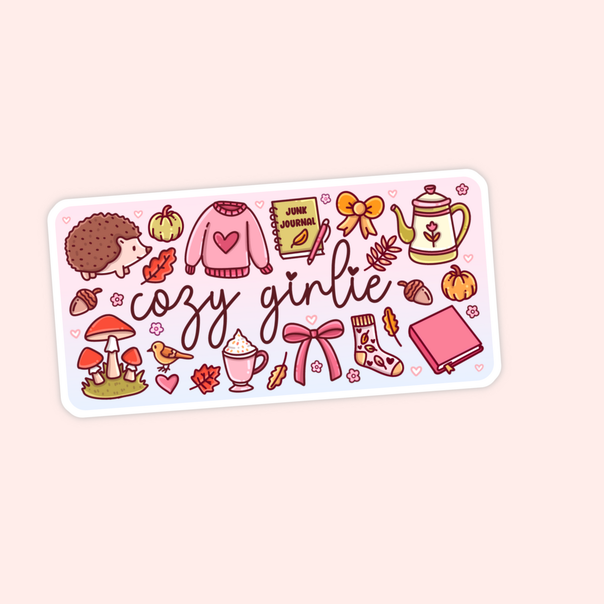A rectangular sticker with rounded corners featuring a cute autumn-themed illustration and the words 'cozy girlie' in cursive. The design includes a hedgehog, sweater, journal, teapot, acorns, mushrooms, a hot cocoa cup, and bows on a pale pink background.