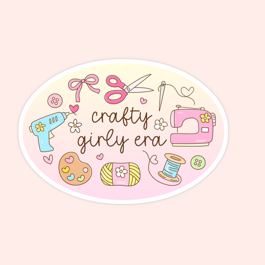 An oval sticker or decal with a white border, featuring cute craft-themed illustrations including a pink sewing machine, scissors, bow, hot glue gun, yarn, spool, and flowers. The center reads 'crafty girly era' in brown script.