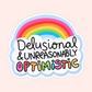 A die-cut sticker featuring a pastel rainbow above the text 'Delusional & Unreasonably Optimistic' in playful handwritten lettering, shown on a pale pink background.