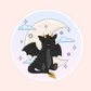 A circular sticker or print featuring a cute black cartoon dragon sitting in front of a large cream-colored crescent moon, with pastel clouds, stars, and a pale pink background.