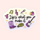 A die-cut sticker with the phrase 'Life's what you make it' in black script, surrounded by colorful doodles including purple cowboy boots, a purple electric guitar, a pink megaphone, a green treble clef, a yellow guitar strap, and small yellow stars on a pale yellow and lavender background. The sticker is centered on a soft pink backdrop.