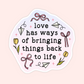 A die-cut sticker with a white border and a light pink background, featuring the quote 'love has ways of bringing things back to life' in black text, surrounded by hand-drawn illustrations of pink ribbons, yellow tulips, paper airplanes, and small pink stars.