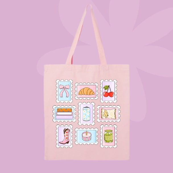 A light pink canvas tote bag with long shoulder straps, featuring a grid of nine cute, vintage-style illustrated postage stamp designs on the front. The designs include a pink bow, a croissant, cherries, a cake, a polka-dot cupcake, a cowboy boot, a jar, a book, and a map, all set against a soft lavender background with subtle leaf silhouettes.
