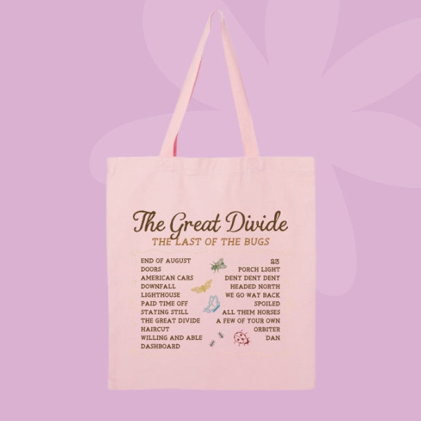 A light pink canvas tote bag with pink handles, featuring a vintage-style printed graphic titled 'The Great Divide: The Last of the Bugs' with a list of bugs and small illustrations, set against a solid lavender-purple background.