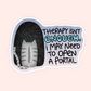 A die-cut sticker featuring a gray tabby cat seen from behind inside a black starry space scene, next to the text 'THERAPY ISN'T ENOUGH. I MAY NEED TO OPEN A PORTAL' in black and light blue lettering.
