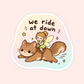 A die-cut sticker featuring a cute cartoon illustration of a brown cat being ridden by a small fairy, with the text 'we ride at dawn' above them. The sticker has a white border and is shown on a soft pastel pink background.