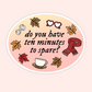 An oval sticker with a pastel gradient background featuring the text 'do you have ten minutes to spare?' in black script. The design includes autumn-themed illustrations such as leaves, a red scarf, heart-shaped glasses, crumpled paper, and a coffee cup on a light pink and peach gradient.