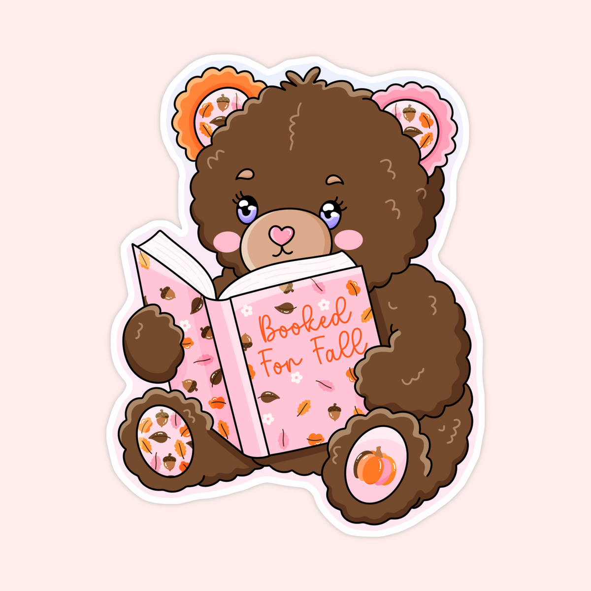 A die-cut sticker featuring a cute cartoon brown teddy bear reading a pink book with autumn leaves and acorns, set against a pale pink background.