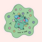A die-cut sticker featuring a cute cartoon frog surrounded by small flowers on a light green background, with the text 'THIS IS FINE' at the bottom, shown on a pale pink background.