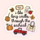 A die-cut sticker with a scalloped edge featuring autumn-themed illustrations and the phrase 'i like long walks through the orchard' in black script. The design includes a caramel apple, pumpkin, red pickup truck, tote bag, apple, admit one ticket, and a carton of apple cider on a pale pink background.