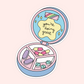 A die-cut sticker featuring a pastel-colored illustrated pill organizer compact with capsules and pills inside, shown open on a light pink background. The inside lid has a yellow cloud shape with the text 'you're doing great' in black script.