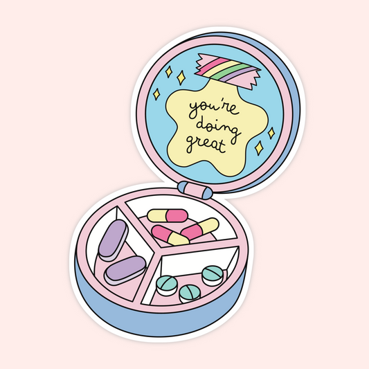 A die-cut sticker featuring a pastel-colored illustrated pill organizer compact with capsules and pills inside, shown open on a light pink background. The inside lid has a yellow cloud shape with the text 'you're doing great' in black script.